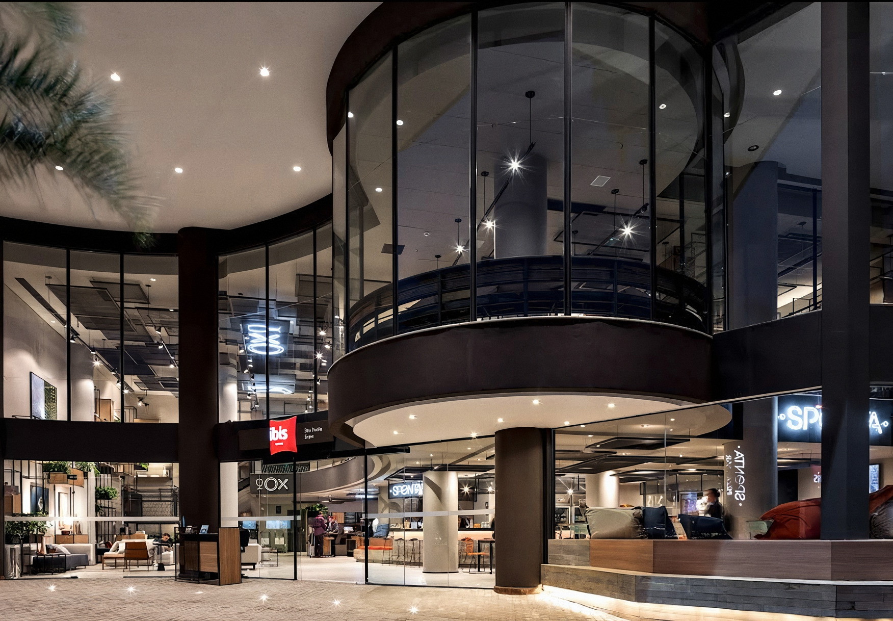 Lobby do hotel Ibis à noite, com pé-direito duplo, fachada de vidro curva e áreas de convivência iluminadas